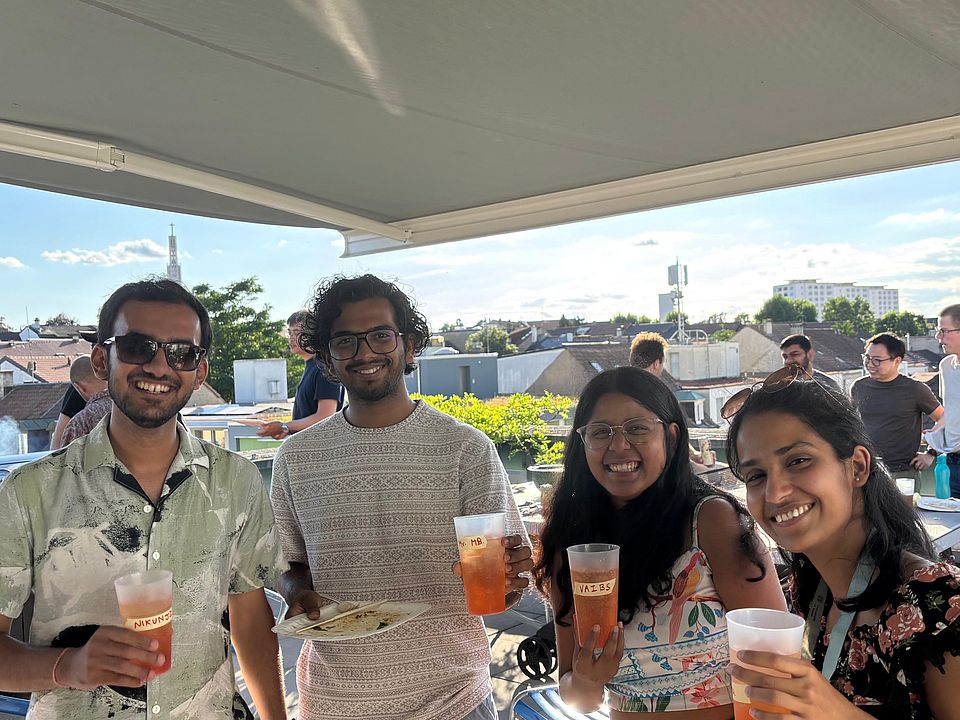 Four smiling PhD students holding drinks and a plate with a crepe, standing on a rooftop terrace with other people and city rooftops in the background.